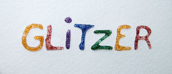 Mit verschiedenfarbigem Glitzerkleber steht auf einem Papier geschrieben: "Glitzer".