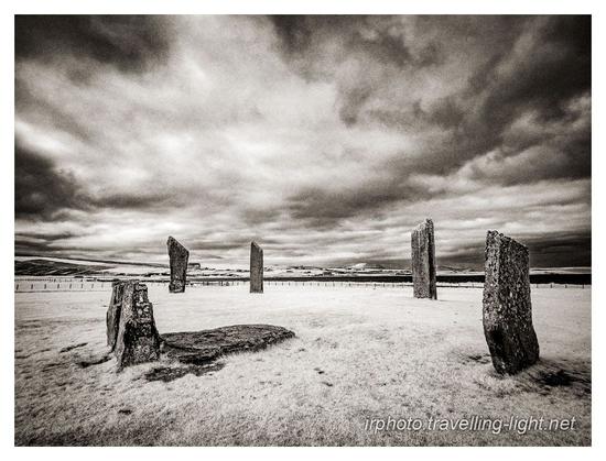 Toned black and white infrared photo of four large flat standing stones in a field with a single recumbent stone and two small uprights, all under a menacing sky.