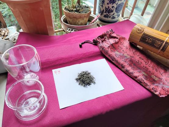 The stage is set.  My omnipresent 茶经 (Classic of Tea) bamboo scroll frames the setting on the right while the glassware I will be brewing the tea in frames it to the left.  The background is the potted plants outside on a sill and underneath it all is the cloth I use to give it a nice colour.  The brocade bag for the scroll sits in front of the scroll, and in the middle is a slip of paper with some tea leaves presented.
