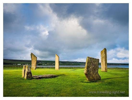 Photo of four large flat standing stones in a field with a single recumbent stone and two small uprights, picked out in a small patch of sunlight, all under a menacing sky.