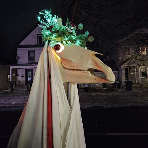 A Mari Lwyd, which is a horse skull decorated with a wreath crown, Christmas ball eyes, and lights, with the back of the skull draped with a sheet and hung with long ribbons.