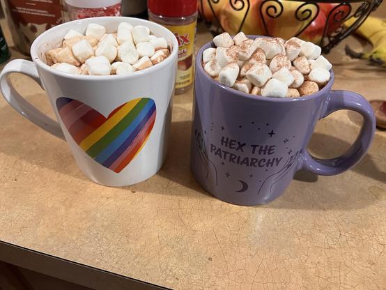 Photo of two mugs overflowing with mini marshmallows. The left mug is decorated with a heart with rainbow stripes across it. The right mug is lavender with “hex the patriarchy” written on it between two hands spreading sparkles.