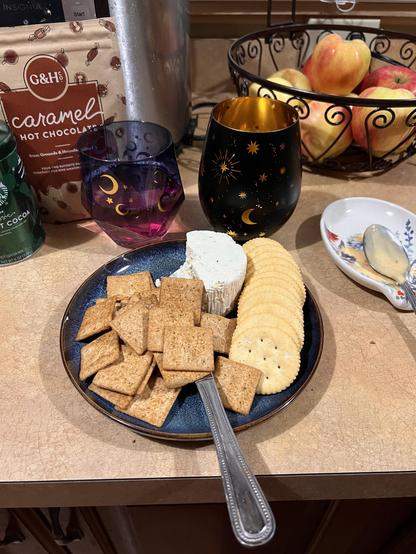Photo of a small blue plate with half of a round herbed cheese, a line of ritz crackers, and a pile of square crackers with herbs on them (they’re tomato basil). In the background are two glasses of wine decorated with the moon, and you can see the hot cocoa packet from earlier