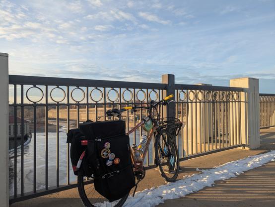 Tan bike with black euphonium case and pannier bag attached to the rear rack on the sidewalk of a bridge. There is a little snow on the deck of the bridge but overall it is exposed concrete.