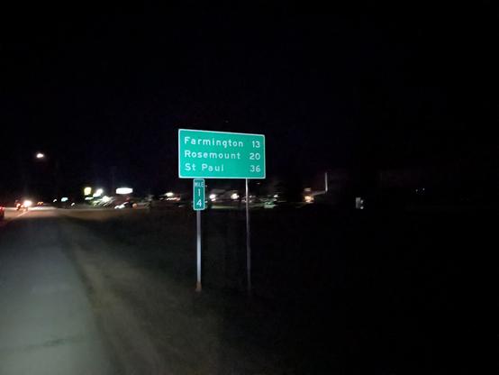 Green sign that says, "Farmington 13, Rosemount 20, St Paul 36" along a road with a 6 foot paved shoulder at night.