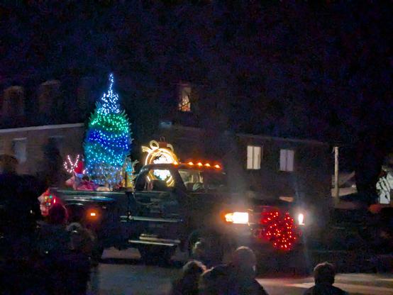 Illuminated tree and light truck in a parade with a lit wreath on the front. It was in front of an apartment building. There are people seated and standing in the foreground.