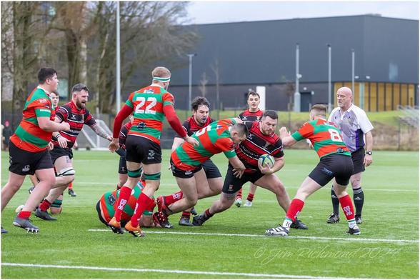 A rugby match in action, featuring players in colorful jerseys. One player is about to be tackled while holding the ball, with others nearby showing varied expressions of effort and focus. An official is observing the play. The scene is set on a plastic pitch.