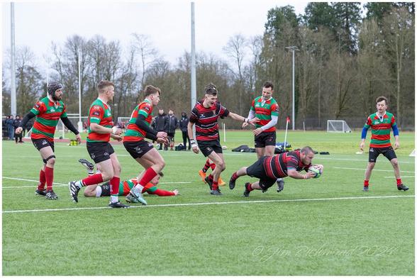 A rugby match on a grass field featuring players in red and green striped jerseys. One player is diving to score, while others are engaged in the play. Spectators are visible in the background.