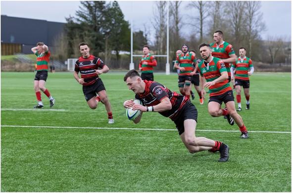 A rugby player in a black and red uniform is diving to score with the ball, while other players in green and red uniforms are in the background. The scene takes place on a plastic rugby pitch.