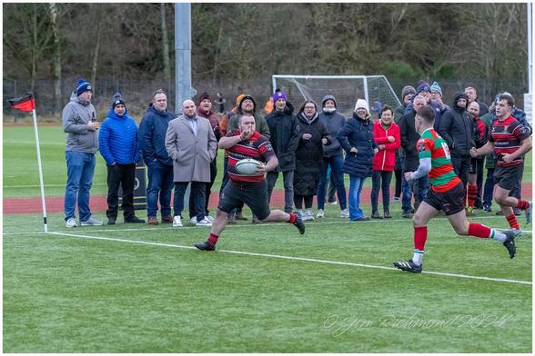 A rugby match in progress, featuring a player in a black and red striped jersey running with a rugby ball. Spectators watch from the sidelines, showing a mix of expressions and attire, against a green field.