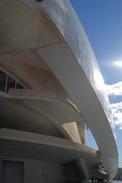 Foto del edificio del palau de les arts de la ciudad de las ciencias de Valencia. El edificio tiene unas formas muy redondeadas y muchas alturas. Tiene una parte interna donde se ve el hormigón y una parte externa blanca como si fuera un casco cubriendo el edificio. La parte externa blanca es un trencadís, un tipo de mosaico a partir de cerámica.