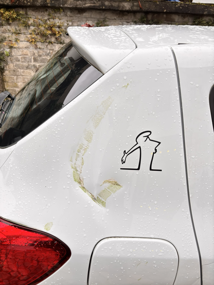 Das Foto zeigt die hintere Seitenpartie eines weißen Autos, das mit Regentropfen bedeckt ist. Auf der lackierten Fläche ist eine deutliche, längliche Beschädigung zu sehen, vermutlich ein Kratzer oder eine Abreibung mit grünlichen Rückständen, die möglicherweise von Pflanzen oder einem anderen Material stammen. Direkt neben der Beschädigung ist ein schwarzer Aufkleber angebracht, der eine stilisierte Figur zeigt: eine gebückte Person mit ausgestrecktem Arm, die etwas vom Boden aufhebt. Im Hintergrund ist eine alte Steinmauer mit vereinzeltem Pflanzenbewuchs zu erkennen. (Text von ChatGPT)