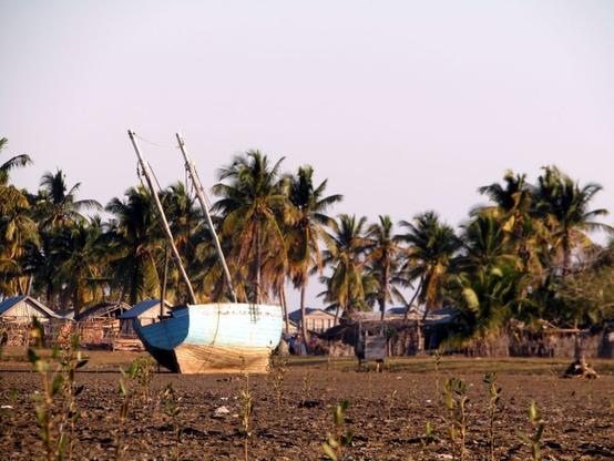 A large Malagasy sailboat rests on the mudflats at low tide, with sparse seagrass scattered in the foreground. In the background, a quiet Malagasy village.
