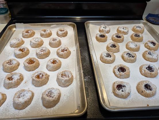 Two baking sheets rest on a stove top. The closest outer corners are cropped due to the photo angle but there's 18 volcano-shaped cookies on each tray. The one on the left has an orange tint in the center of each cookie as a round gem of baked apricot jam, and the tray on the right has the same but a deep dark rich red from the baked raspberry jam. All the cookies are dusted with powdered sugar, like a light snowfall.