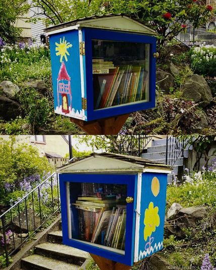 A standard Little Free Library box is painted a bright blue and has a couple of scenes obviously painted by small children: on one side is a red house under a bright yellow dandelion sun in a blue sky, on the other side is a yellow flower in grass under a yellow sun