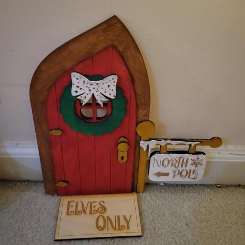 Painted wood door with wreath and snow. A sign says "North Pole" and the mat says "elves only"