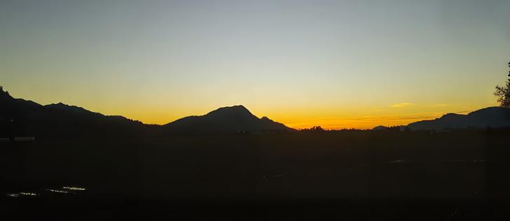 A serene twilight landscape featuring a silhouette of mountain peaks against a gradient sky transitioning from deep blue at the top to warm yellow and orange hues near the horizon. The sun has set, leaving a tranquil glow over the valley. Dark, open land stretches below the mountains, dotted with faint reflections of water in the distance. Sparse trees and vegetation frame the edges, enhancing the natural stillness of the scene.