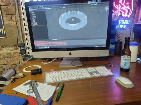 Ein Computerbildschirm auf einem Schreibtisch mit Teilen, Werkzeug und Messwerkzeug zeigt ein CAD Programm mit einem Zahnrad. Neben der Tastatur steht eine Flasche alkoholfreies Bier. An der Wand hängt ein Leuchtwerbeschild mit einem rosa Elefanten.