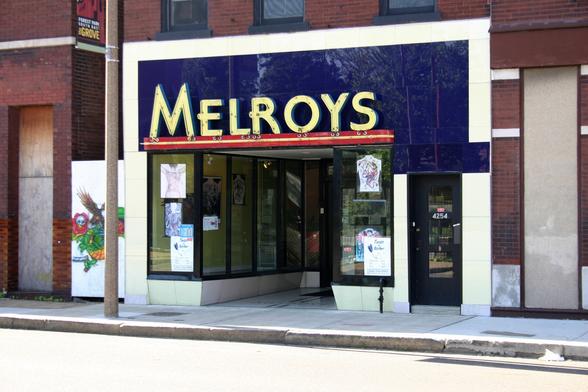 Color photo of an Art Deco-style storefront, with a sign in sans-serif Art Deco letters saying "Melroys". The letters are yellow, on a dark blue background, with a red-yellow-red striped band below.