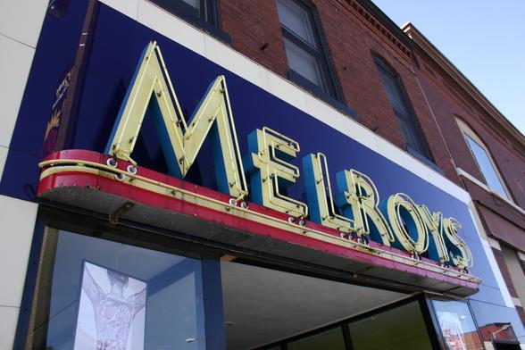 Color photo of an Art Deco-style storefront, with a sign in sans-serif Art Deco letters saying "Melroys". The letters are yellow, on a dark