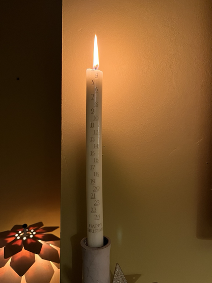 A tall, slender, cream coloured advent candle is burning. There are gold coloured numbers from 4 down to 24 printed on to the candle (indicating that 1 through to 4 have been burned through), with HAPPY CHRISTMAS printed in gold at the bottom of the candle