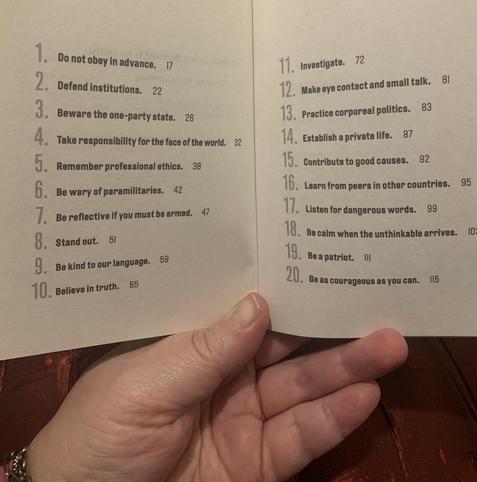 A hand holds up a numberedTable of Contents from inside the book “On Tyranny.” 1: Do not obey in advance. 2: Defend institutions. 3: Beware the one-party state. 4: Take responsibility for the face of the world. 5: Remember professional ethics. 6: Be wary of paramilitaries. 7: Be reflective if you must be armed. 8: Stand out. 9: Be kind to our language. 10: Believe in truth. 11: Investigate. 12: Make eye contact and small talk. 13: Practice corporeal politics. 14: Establish a private life. 15: Contribute to good causes. 16: Learn from peers in other countries. 17: Listen for dangerous words. 18: Be calm when the unthinkable arrives. 19: Be a patriot. 20: Be as courageous as you can.