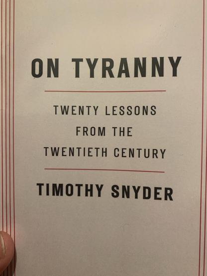 A hand holds up a paperback front cover that reads: ON TYRANNY TWENTY LESSONS FROM THE TWENTIETH CENTURY (by) TIMOTHY SNYDER