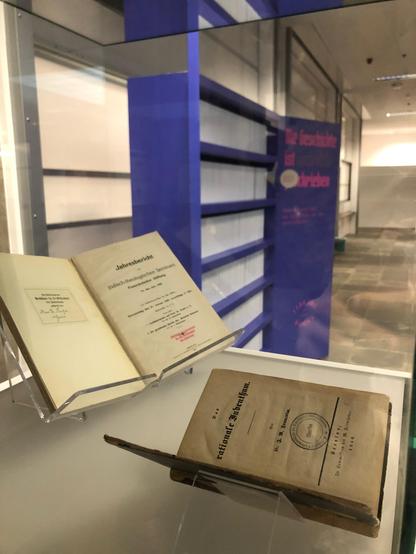 Zwei Bücher liegen in einer Vitrine. Dahinter ist das blaue Ausstellungsmodul in Regalform des Projekt "Library of Lost Books" zu sehen