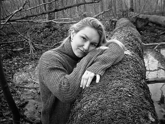 A young blonde woman wearing an oversized sweater leans against the side of a fallen tree in black and white.