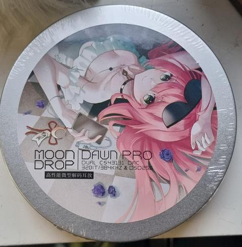 A photo of a round aluminum tin with a round sticker on it featuring a pink haired anime girl laying on the floor, on her back, listening to music on her phone using headphones and a USB-C amp. The phone has a cresent moon charm. There's little purple roses all over the floor and at the bottom it says "MOON DROP | DAWN PRO DUAL C543131 DAC 32 BIT / 384 KHZ & D5D256". There's also some text in Chinese below that, but I don't know what it says.