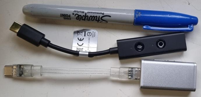 A photo of a blue sharpie permanent marker, a black soundblaster USB-C amp, and a silver Moondrop USB-c amp. The sharpie is maybe a cm shorter is maybe a cm shorter than the soundblaster amp, and 2cm shorter than the moondrop amp.

This photo shows the front of both of the amps, unlike the last photo.

The soundblaster amp has two buttons, a mute button, and another button with a circle that I'm not sure what it does.

The moondrop amp is smooth with no buttons or holes on the top, but you can see the side volume buttons slightly peaking out.