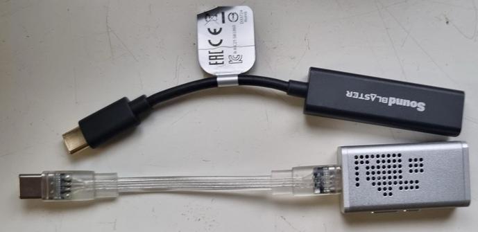 A photo of two USB-C amps side by side upside down on a white surface. The top one is a black soundblaster amp and the cable is slightly curved with heavy use. The amp part is probably about 3cm-sh long, 1.5cm wide, and maybe .5cm thick. The 4cmish long cable part has a tag with a bunch of symbols on it for it's rating (I don't know what those mean).

Below it is a silver amp with lots of little holes in it (I guess for venilation?). It's thicker (1cm-ish) and wider (2cm-ish), but shorter (2cm-ish). The cable coming out of it is clear transparent, detachable, and USB-C on both ends.