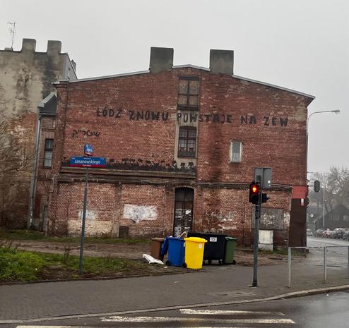 Zdjęcie przedstawia stary i pomazany budynek z czerwonej cegły, stojący przy skrzyżowaniu ulic Zgierskiej i Limanowskiego w Łodzi. Jest brzydka pogoda. Przed budynkiem widać kolorowe kosze na śmieci. Na budynku ktoś zostawił czarny napis: „ŁÓDŹ ZNOWU POWSTAJE NA ZEW”