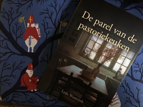Boekomslag van de roman De parel van de pastoriekeuken. Een keuken met daarin een flinke houten tafel en stoelen er omheen. In de vensterbank een Mariabeeld.