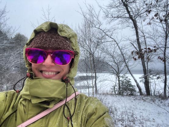 Picture of Joanne Merriam bundled up in layers of winter coat and hat smiling into the camera with snow-covered trees and lake behind her.