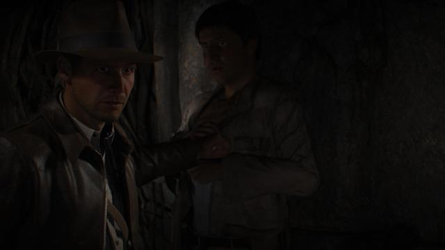 In a dimly lit cave, Indiana Jones pushes his companion against a wall to stop him from triggering a trap.