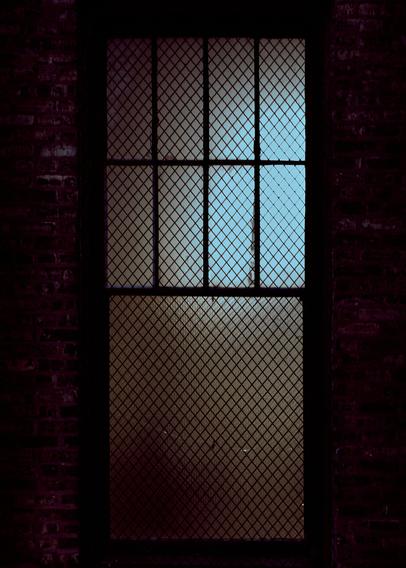 A barred window with a crisscross metal grill, set in a brick wall. The window is partly illuminated with a blue hue at dusk, creating a dim and moody appearance.
