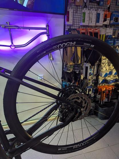 Заднее колесо велосипеда с покрышкой Schwalbe G-One Allround и ободом DT Swiss R500db, установленного на ремонтной стойке в веломагазине. На заднем плане видны различные велосипедные аксессуары на витрине, подсвеченной фиолетовой подсветкой.