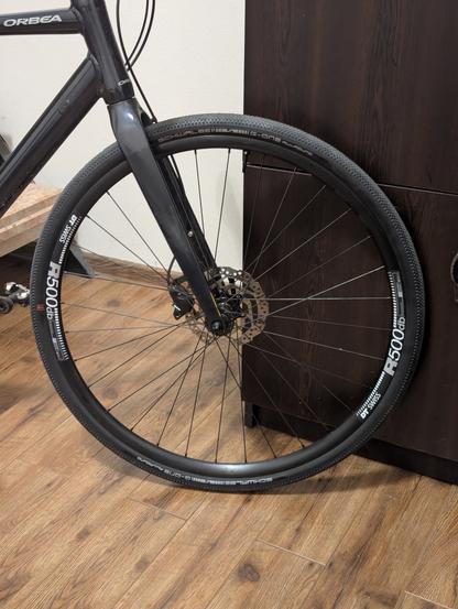 Переднее колесо велосипеда с покрышкой Schwalbe G-One Allround и ободом DT Swiss R500db, установленное на велосипеде марки Orbea. Велосипед стоит на деревянном полу рядом с темной мебелью.