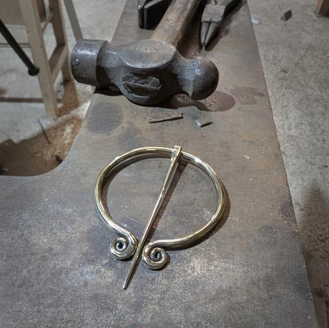 Une grande fibule en laiton (8cm de diamètre) avec ses deux extrémités spiralées, posée sur la table d'une grosse enclume, on voit quelques outils en haut du cadre (marteau, pinces)