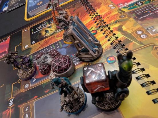 Close-up of game board. Several minis, some out of focus, representing player characters, an outpost, a launchpad, and an alpaca carrying a red die.