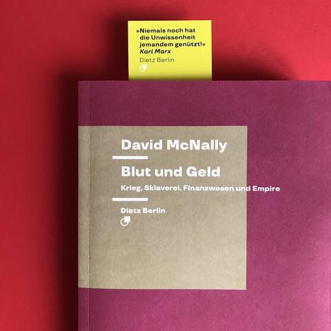 Cover des Buches »Blut und Geld. Krieg, Sklaverei, Finanzwesen und Empire« von David McNally. Darin steckt ein Dietz-Lesezeichen mit dem Aufdruck: „Niemals noch hat die Unwissenheit jemandem genützt!”, Zitat von Karl Marx