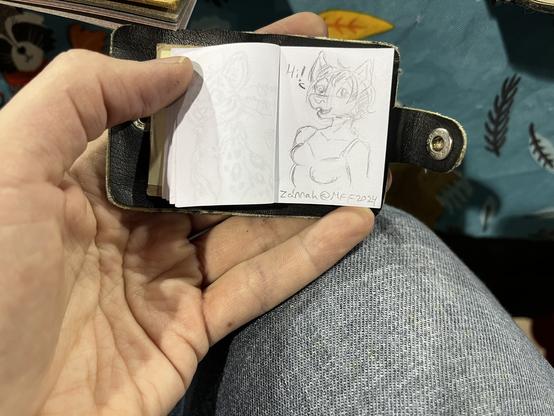 The tiniest sketchbook commission ever. A 1”x1.5” book in a white person’s hand with. Little sketch of a fox girl from bust up saying hi