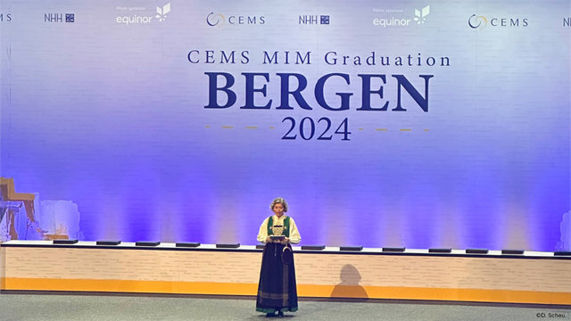 A woman in a Norwegian-style costume stands on a stage and reads something aloud. Behind her a projection of the following text: CEMS MIM Graduation - BERGEN - 2024