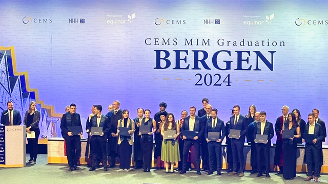 A group of CEMS MIM graduates stand for a photo with stylish certificates on stage in front of a large audience at the CEMS MIM Graduation 2024 in Bergen.