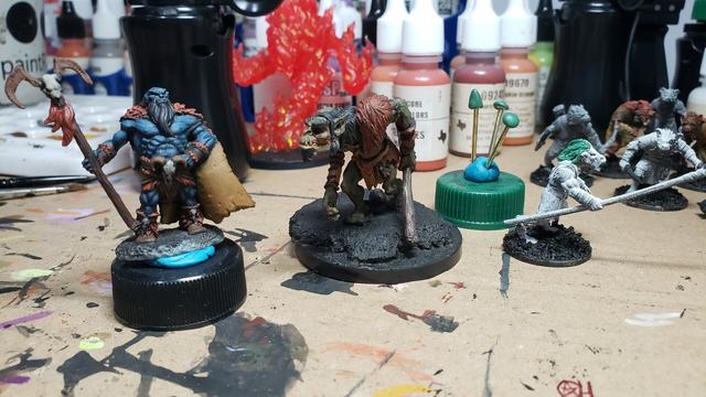 Buff Orc sorcerer with skull headed staff on the left. He's painted with blue skin wearing orange red fur cloak, boots, wrap, etc. 
I'm the middle is an old metal troll with a club. 
To the right is a Frostgrave gnoll (unpainted) with a GW gor head and greenstuff hair. Next to it is a bottle cap with 3 nails in it. Each mail has a greenstuff mushroom cap sculpted on it
