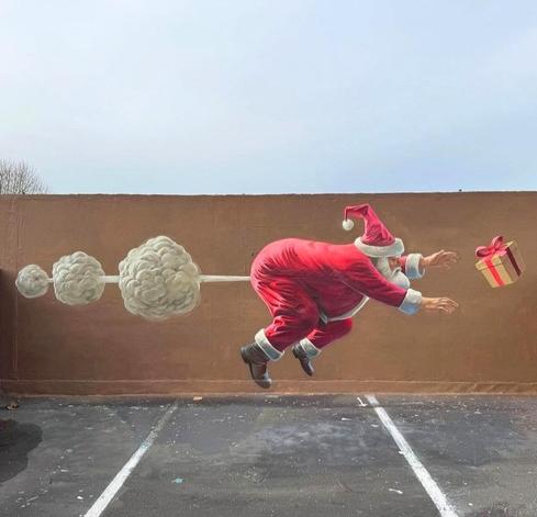 Streetartwall. On a long brown wall, at the top of a parking deck, there is a funny mural of a cartoon-style Santa Claus who is struggling with small problems. The fat, red Santa is flying over the wall trying to reach a gift package flying in the air in front of him. He has farted and the double wind backlash seems to have thrown him off course. Sorry for the alt text, but that's exactly what was painted on the wall.