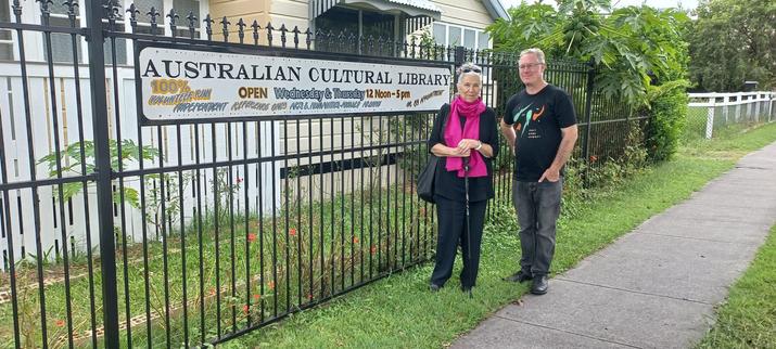 A couple of volunteers standing outside the Australian Cultural Library in front of the sign, showing it's open Wed & Thu 12-5pm, or by appointment.