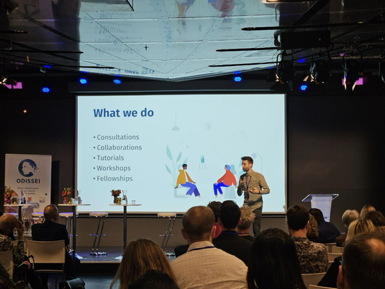 Speaker Erik-Jan van Kesteren standing on stage in front of a crowd, behind him a presentation slide that reads: what we do; consultations, collaborations, tutorials, workshops, fellowships