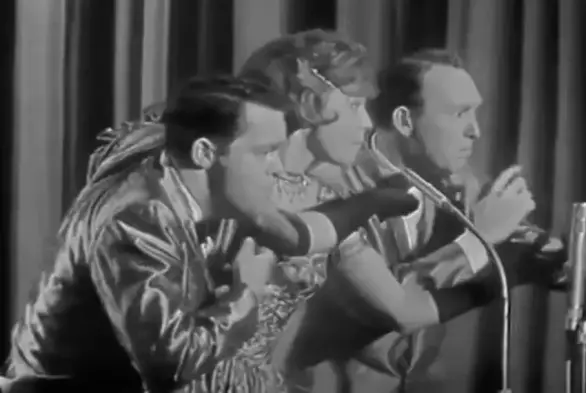 A black-and-white screenshot of one of the clips. All three (from left: Svend, Alice, and Ulrik) are leaning toward their microphones. They’re dressed in shiny show attire, their faces show concentration with a hint of comedic attitude.